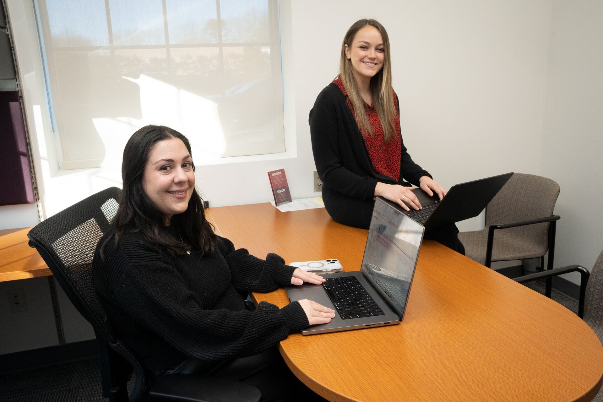 Gina Desio and Shannon Seymour smiling at the camera while working on their computers