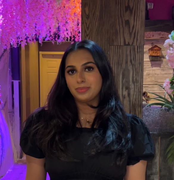 Person with long dark hair wearing a black top, standing in front of a colorful background with purple foliage and a multi-colored doorway