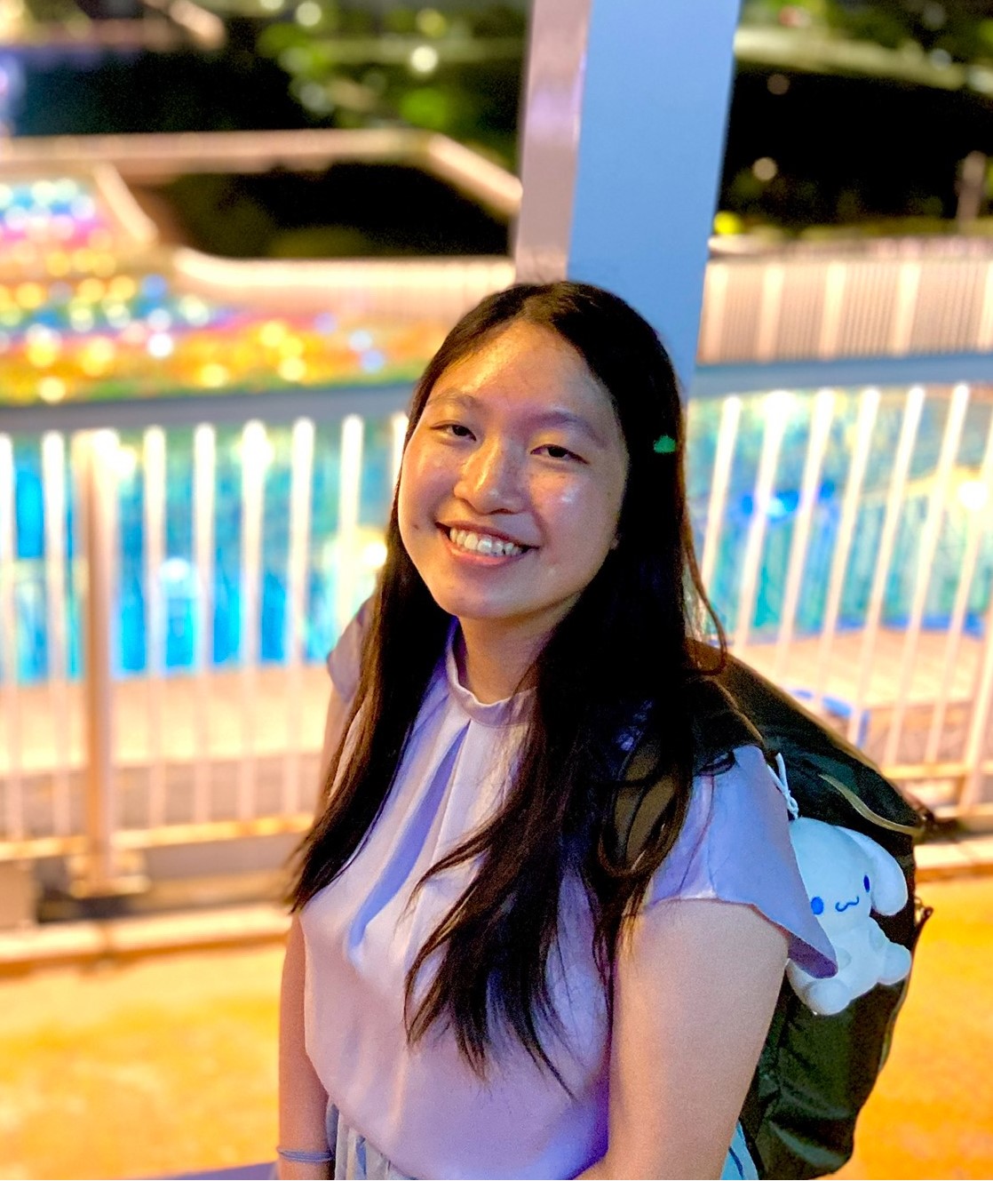 A person smiling, standing in front of illuminated railings at night, wearing a backpack with a plush keychain.