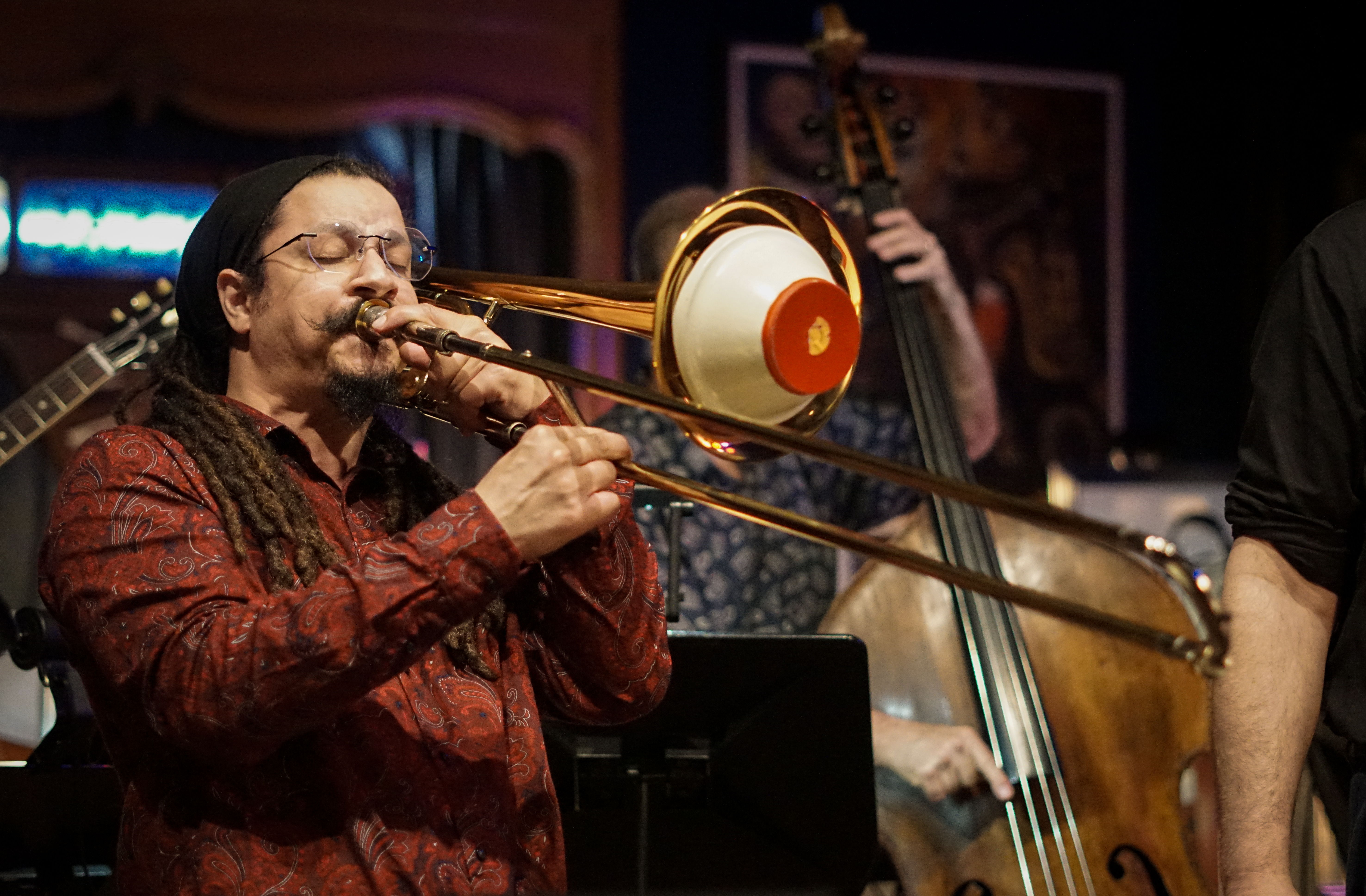 A musician playing a trombone on stage with a double bass player in the background, in a live jazz performance setting.