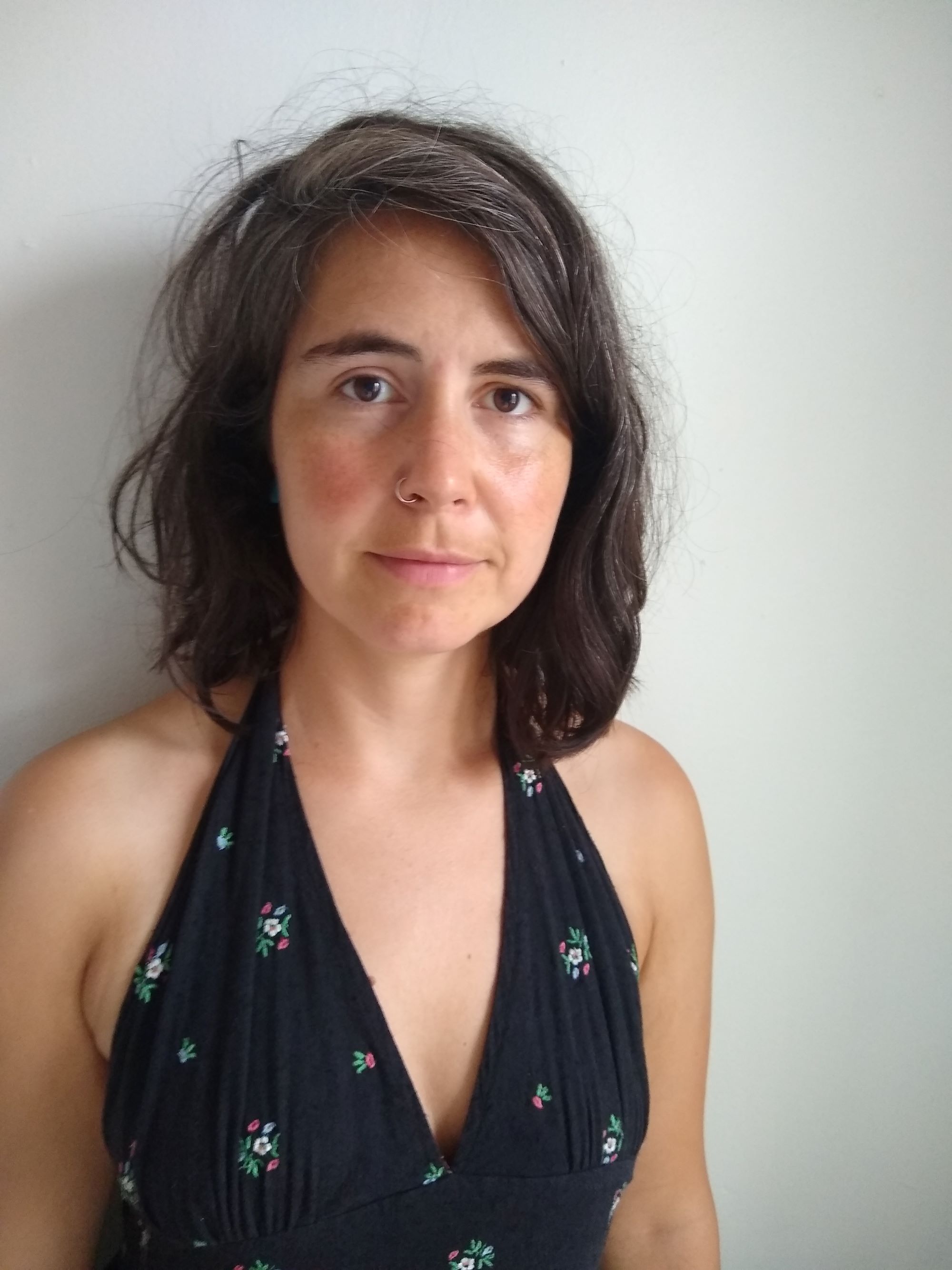 A person with shoulder-length brown hair, wearing a black floral top, standing in front of a white wall and looking directly at the camera.