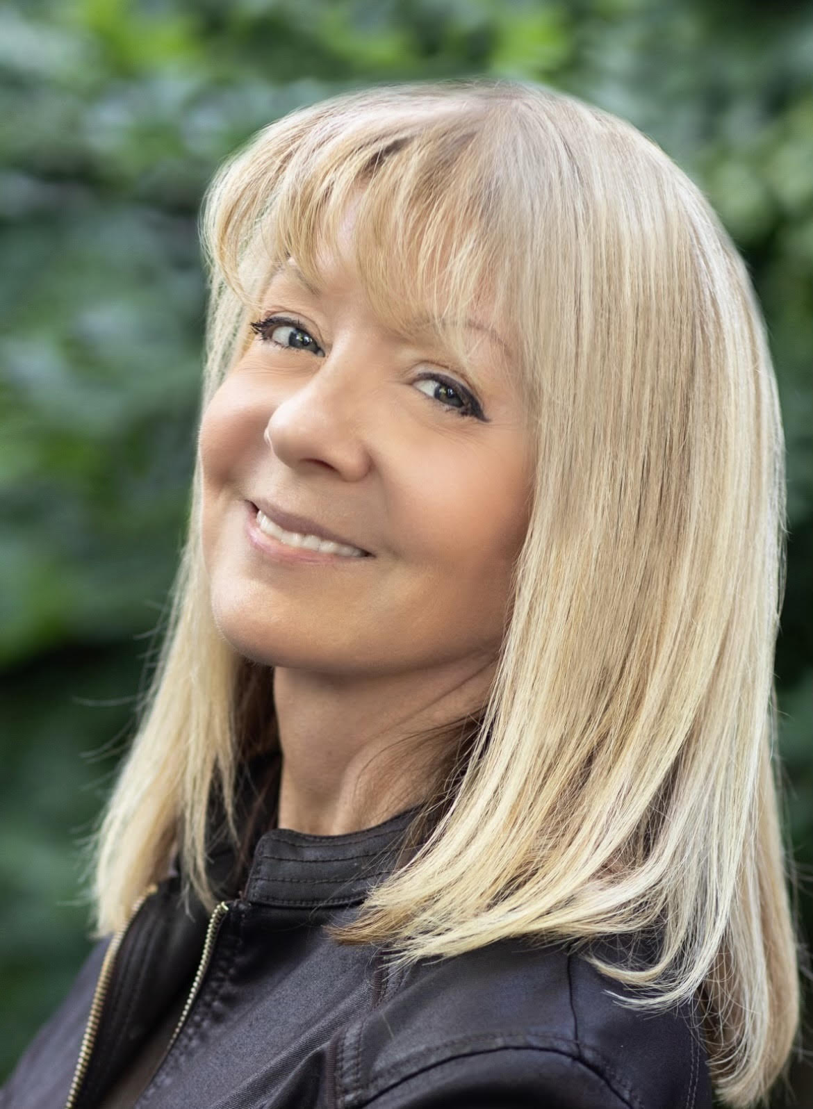 Woman smiling in a set against a blurred green background. She has long blonde hair and is wearing a black leather jacket.