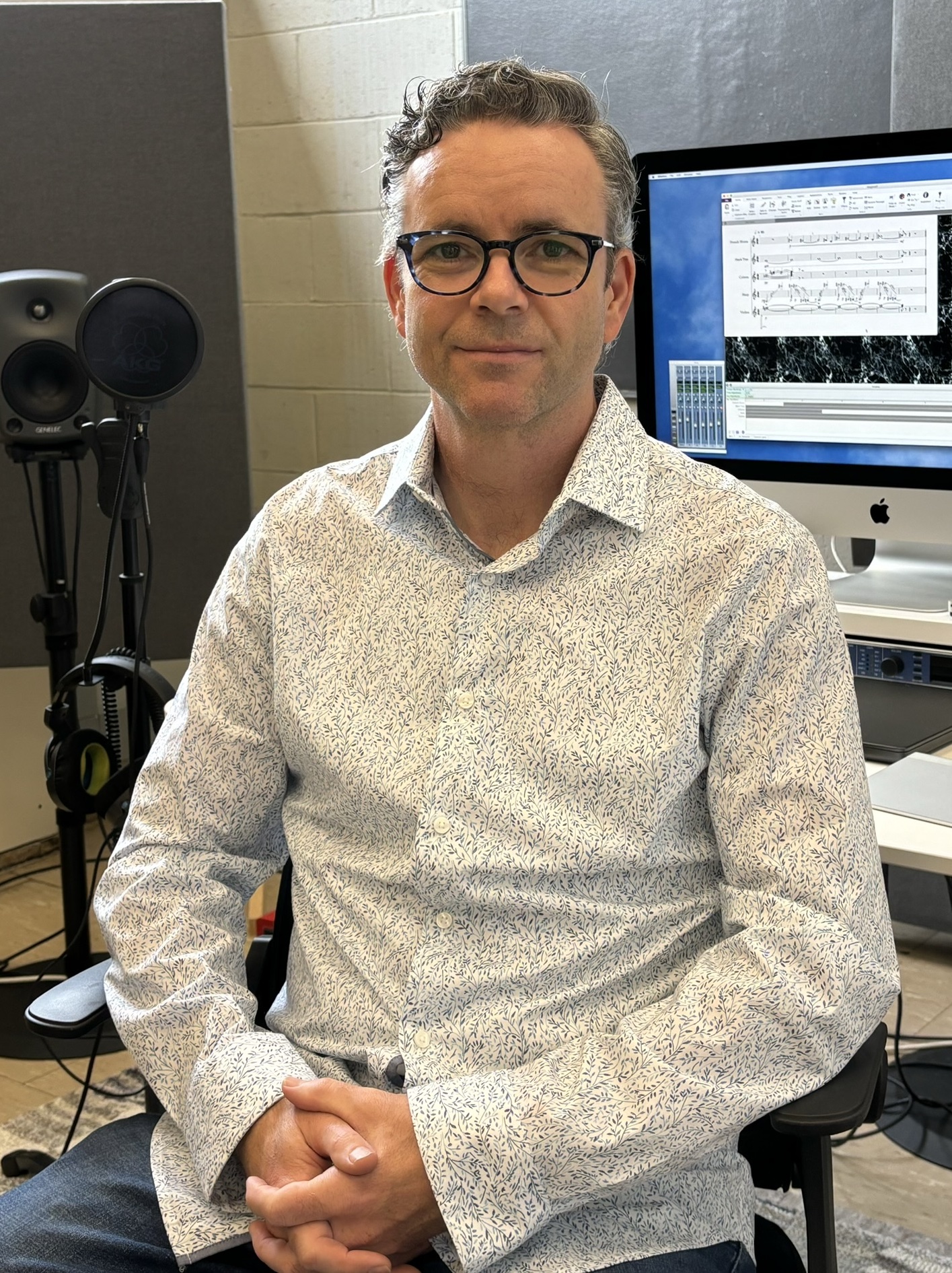 portrait of Tony Doyle, with a slight smile, sitting down and looking forward, wearing a white shirt with blue pattern and light blue jeans.