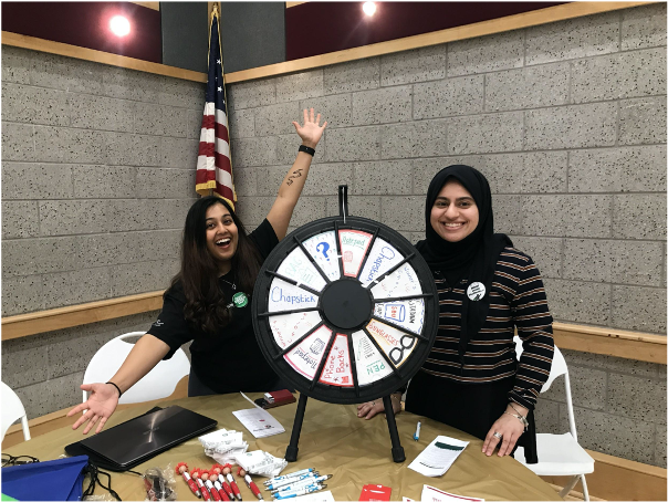 two peer educators standing behind a table with a trivia wheel on the table. Both students look excited. 